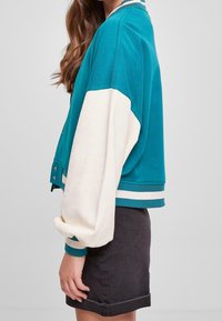 Kobieta w turkusowej i białej kurtce z dużymi rękawami, nosząca czarną spódnicę, stojąca w profilu na jednolitym tle.