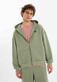 Ljusgrön zip-up hoodie av mjukt tyg, med en framficka, sänkt axel och en liten svart logotyp på bröstet.