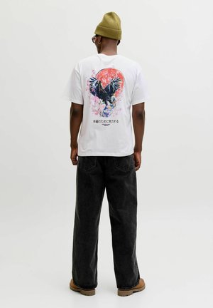 Man draagt een wit T-shirt met een adelaar-afbeelding en een rood zonontwerp, zwarte jeans met wijde pijpen, een mosterdgele muts en bruine schoenen, met zijn rug naar de camera.