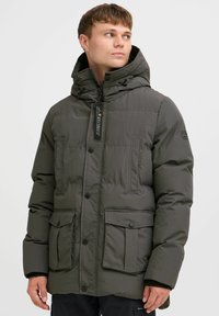Chaqueta acolchada de color verde oscuro con capucha, que cuenta con cierres de cremallera y botón, dos bolsillos frontales y una tela de nailon texturizada.