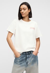 Γυναίκα με κοντά σκούρα μαλλιά, που φοράει λευκό T-shirt Diesel και ξεθωριασμένο μπλε τζιν, με τα χέρια στις τσέπες, κοιτάζοντας ελαφρώς προς τα κάτω και στο πλάι.