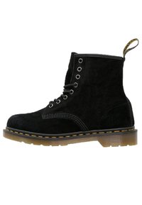 Botín de ante negro con cordones, costuras amarillas, suela translúcida marrón y tira de tela en la parte trasera.