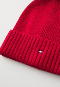 Rød ribbestrikket lue med brettet oppkant. Har en liten logo med blått, hvitt og rødt på oppkanten. Myk tekstur, avslappet design.