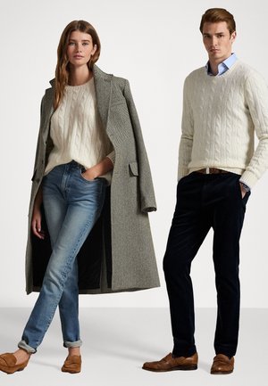 Twee modellen dragen gebreide truien, een visgraatjas, denim en donkere broeken. De jas is grijs en beide truien zijn crèmekleurig. De schoenen zijn bruin.