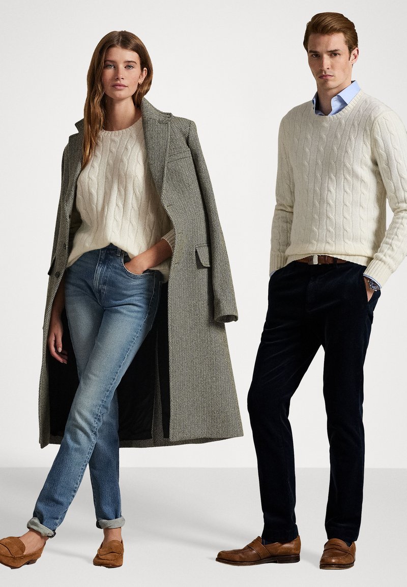 Deux modèles portent des pulls en tricot, un manteau en chevrons, du denim et un pantalon foncé. Le manteau est gris et les deux pulls sont couleur crème. Les chaussures sont marron.