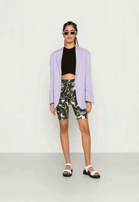 Blazer oversized lilás, top cropped preto, calções de bicicleta com padrão, sandálias brancas e óculos de sol. Design moderno com texturas contrastantes.