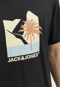 Camiseta de algodón negra con un diseño gráfico que incluye una palmera, una montaña y agua en beige y azul claro, con el texto "JACK&JONES" en blanco.