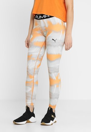 Leggings avec un motif abstrait orange et gris, fabriqués en tissu extensible. Logo PUMA noir sur la taille, associés à des chaussures de sport noires.