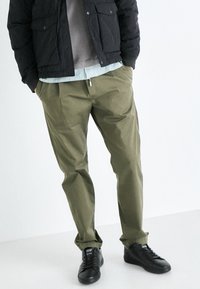 Pantalones verdes oliva de corte relajado, combinados con una chaqueta acolchada negra y zapatillas negras. Lleva una camisa a rayas debajo.