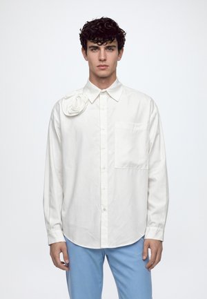 Un homme porte une chemise blanche boutonnée, ornée d’une rose en tissu sur l’épaule et dotée d’une poche poitrine, avec un pantalon bleu clair.