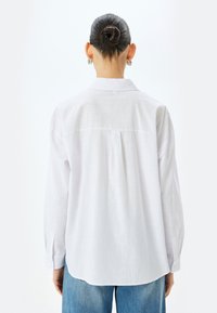 Camicia bianca a maniche lunghe con colletto, dettaglio delle pieghe posteriori e polsini con bottoni, realizzata in un tessuto leggero e testurizzato. Abbinata a jeans in denim blu.
