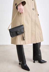Bolso cruzado de cuero negro con acabado texturizado, correa de cadena dorada y detalle de cierre con logotipo, sostenido por una mano que lleva un abrigo beige.