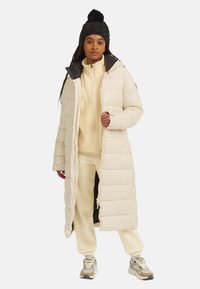 Langer, beiger Daunenmantel mit horizontalem Steppdesign, hohem Kragen und verdecktem Reißverschluss. Getragen über einem cremefarbenen Fleece-Outfit und Sneakers.