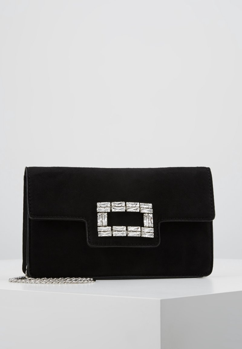 Clutch de ante negro con hebilla rectangular de pedrería y correa de cadena plateada, descansando sobre una superficie blanca.
