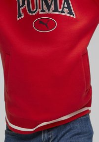 Sweatshirt rouge avec un grand logo "PUMA" et un petit graphique de puma. Dispose de poignets côtelés et d'une bande blanche en bas. Matériau doux.