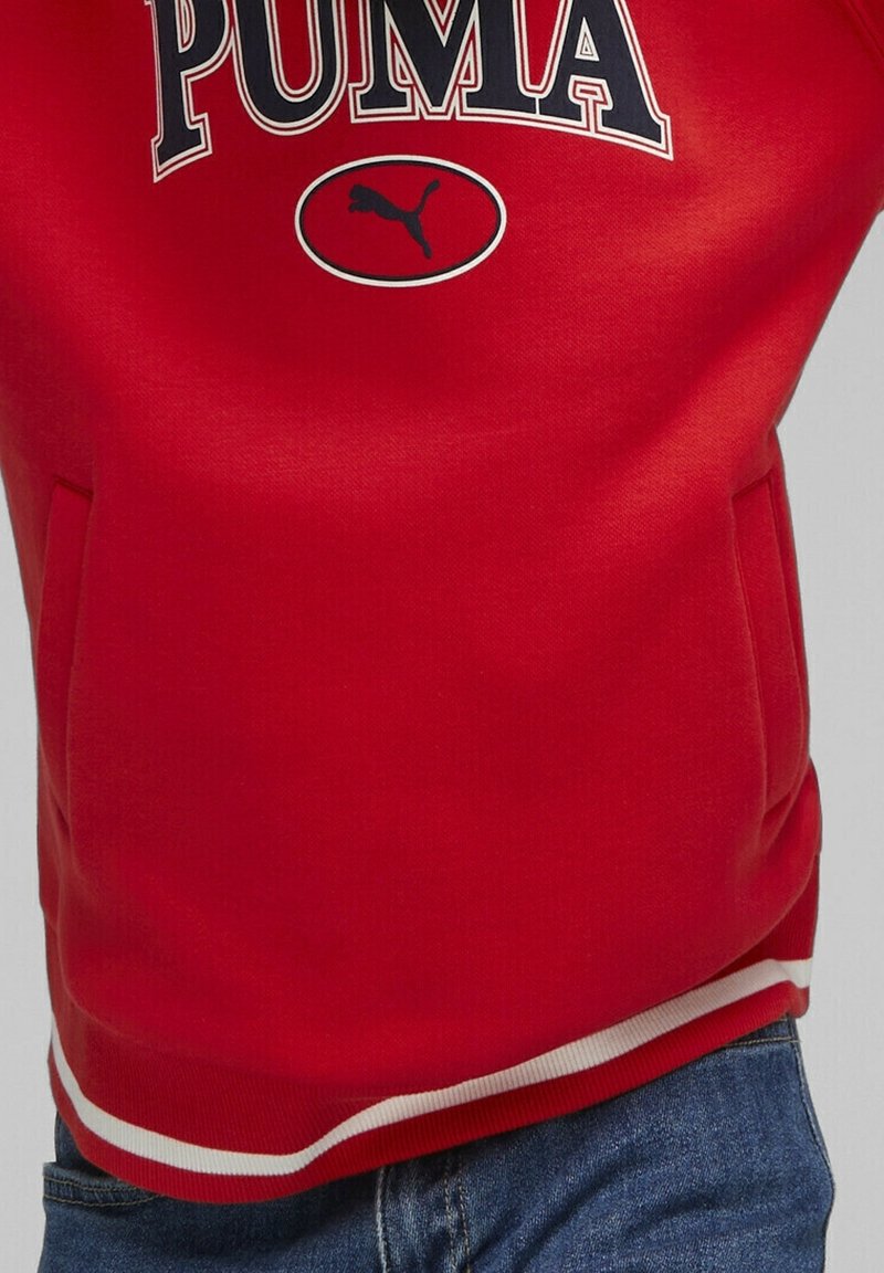 Sweatshirt rouge avec un grand logo "PUMA" et un petit graphique de puma. Dispose de poignets côtelés et d'une bande blanche en bas. Matériau doux.