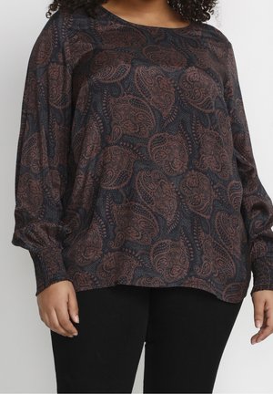Mujer con una camiseta de manga larga oscura con patrón paisley marrón y pantalones negros, mostrada desde el cuello hasta las caderas contra un fondo liso.