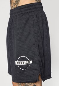 Shorts en maille noire avec une taille élastique, des poches latérales et un logo "CELTICS" blanc en bas à gauche. Présente un design à ourlet fendu.