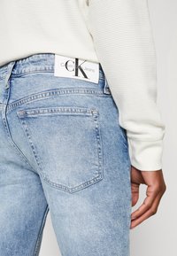 Ljust blå denimjeans med en klassisk femficksdesign, med en vit "Calvin Klein Jeans"-tagg på midjebandet bak.