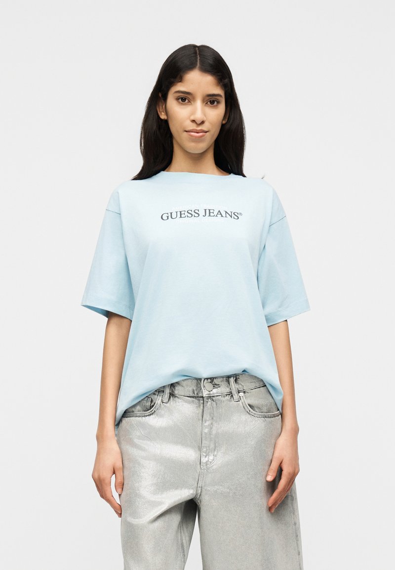 Νεαρή γυναίκα που φορά ένα ανοιχτό μπλε φαρδύ T-shirt με το κείμενο "GUESS JEANS" και μεταλλικά ασημί τζιν ψηλόμεσα, στέκεται μπροστά σε λευκό φόντο.