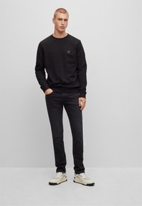 Felpa nera a girocollo con tessuto testurizzato, jeans neri slim e sneakers bianche. La felpa presenta un piccolo logo ricamato sul petto.