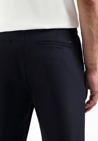 Navyblauwe broek van een gladde stof, met twee achterzakken en subtiele stiksels. Een klein logo is zichtbaar nabij de zak.