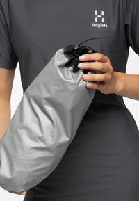 Grå, mjuk, vattentät väska med dragsko, hålls av en person i en mörkgrå skjorta, med en slät yta och minimalistisk design.