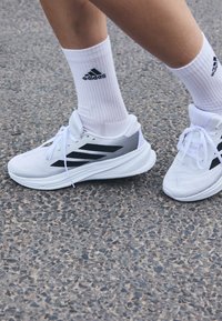 Chaussures de sport blanches avec des accents noirs, tige en mesh et lacets plats ; portées avec des chaussettes blanches à côtes avec un logo noir.