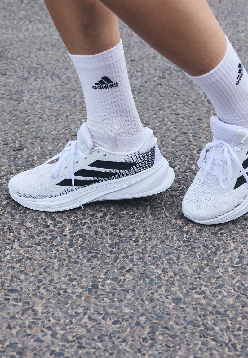 Chaussures de sport blanches avec des accents noirs, tige en mesh et lacets plats ; portées avec des chaussettes blanches à côtes avec un logo noir.