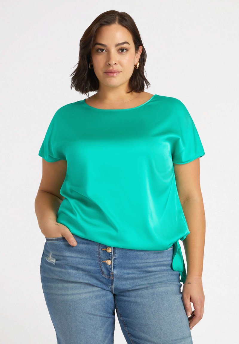 Tealfärgad kortärmad blus i mjukt tyg, med rundad halsringning och subtila sidoknytningar. Parat med ljusblå slitna jeans.