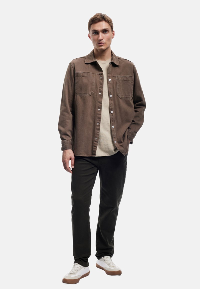 Bruine blouse met knopen en twee borstzakken, gedragen over een lichtbeige top, gecombineerd met donkergroene broeken en witte sneakers met bruin accent.