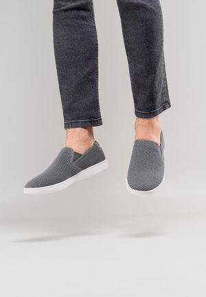 Benen met donkere jeans en grijze slip-on mesh sneakers, licht verhoogd tegen een lichtgrijze achtergrond.