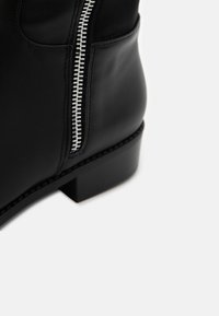 Svart lær ankelstøvel med glidelås på siden, flatt hæl og sømde detaljer langs sålen. Glatt tekstur med et slankt design.