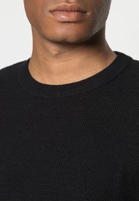 Pull noir en maille avec un col rond côtelé. Vue rapprochée mettant en avant la texture et les détails du tissu.