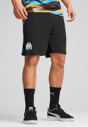 Homme portant un short de sport noir avec un logo blanc Olympique de Marseille, des chaussettes noires Puma et des baskets noires Puma sur un fond gris.