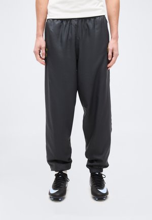 Personne portant un pantalon de sport noir ample et des chaussures à crampons noires avec des logos Nike bleu clair, debout devant un fond uni.
