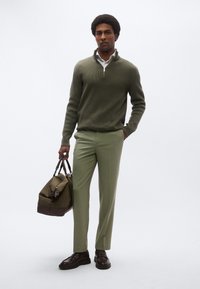 Olivengrøn striktrøje med halv lynlås, lysegrønne skræddersyede bukser og mørke lædersko. Bærer en lærredstaske med læderdetaljer.