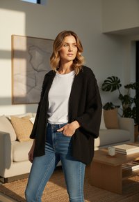 Czarny dziergany kardigan z szerokimi rękawami, noszony na białej koszuli, zestawiony z niebieskimi dżinsami; miękka tekstura, luźny krój i otwarty przód.