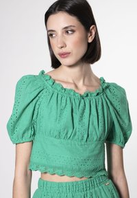 Top corto verde in tessuto testurizzato con maniche a sbuffo, bordo in pizzo e dettagli ad occhiello. Abbinato a una gonna coordinata.