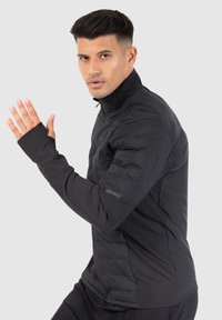 Veste ajustée noire avec un col haut et des fentes pour les pouces. Présente une texture matelassée sur le haut du corps et un tissu lisse sur la partie inférieure.