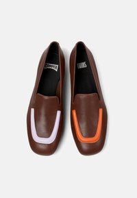 Zapatos de piel marrón tipo slip-on con acentos contrastantes: un diseño contorneado en púrpura claro y naranja en la parte delantera. Textura suave, forma redondeada.