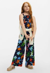 Bloemige jumpsuit met een gerimpelde bodice in marineblauw, met levendige kleuren. Gecombineerd met een crèmekleurige trui die om de taille is geknoopt en geweven sandaaltjes.