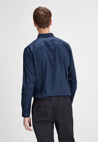 Granatowa koszula z długim rękawem o gładkiej fakturze, z mankietami na guziki i karczkiem na plecach. Połączona z czarnymi spodniami z tylnymi kieszeniami.