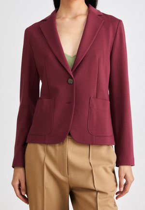 Vrouw draagt een op maat gemaakte bordeauxrode blazer met voorzakken, enkele knoopsluiting, over een groene top, gecombineerd met beige hooggetailleerde broek.