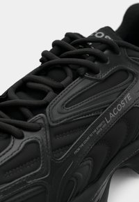 Gros plan sur une basket noire Lacoste avec des lacets épais, des empiècements texturés, et le texte « From the court to the streets » sur le panneau latéral.