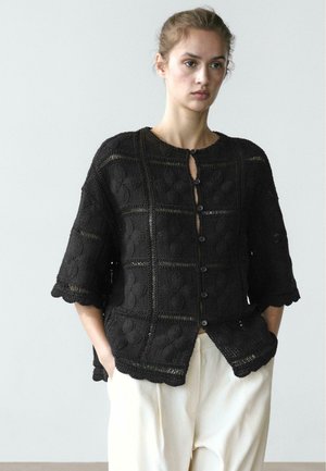 Giovane donna che indossa un cardigan nero lavorato a uncinetto con bottoni e motivi floreali, pantaloni ampi color crema, mani nelle tasche, sfondo neutro.