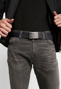Ceinture tissée bleue marine avec boucle en métal argenté, arborant un logo. Associée à un jean gris foncé et à une chemise noire sous une veste noire.