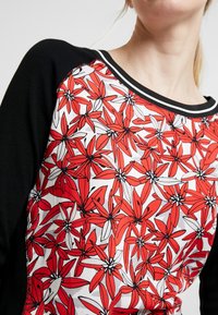 Vrouw met een zwart-mouwen shirt met een rood en witte bloemenpatroon aan de voorkant en een ronde halslijn met witte rand.