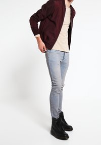 Burgunderfarbene Bomberjacke über einem beigen Hemd, kombiniert mit hellblauen Skinny-Jeans und schwarzen Schnürkampfstiefeln. Glatter Stoff und figurbetontes Design.
