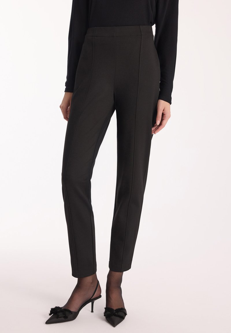 Czarne spodni szyte na miarę z wysokim stanem, z pionowymi szwami i dopasowanym krojem. Zestawione z czarnymi czółenkami na odsłoniętej pięcie z dekoracyjnymi akcentami.